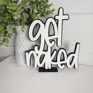 Peut inclure: Panneau en bois blanc avec l'inscription "get naked" en police cursive avec un contour noir. Le panneau est posé sur une base noire. Un vase blanc avec de la verdure est en arrière-plan.