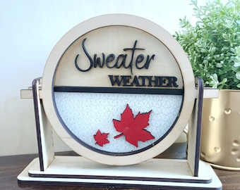 Puede incluir: Un letrero de madera con un fondo redondo blanco y un borde negro. El letrero dice "Sweater Weather" en letras negras de escritura cursiva. Hay dos hojas de arce rojas en el letrero. El letrero está en un soporte de madera.