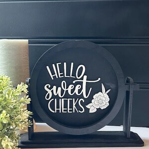 May include: Black wooden sign with the text "Hello sweet cheeks" in white lettering and a white floral design. The sign is on a black stand.