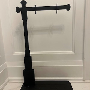 Puede incluir: Soporte de metal negro con una barra horizontal y una base redonda. El soporte está diseñado para sostener artículos como joyas u otros objetos pequeños.