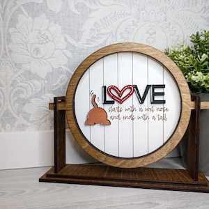 May include: A round wooden sign with the word "LOVE" in black letters and a red heart in the middle. The sign also features a brown cartoon cat with a tail and the text "starts with a wet nose and ends with a tail".