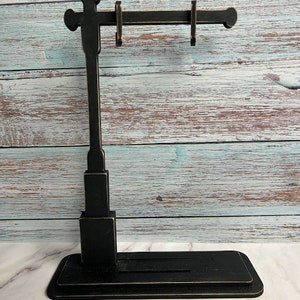 May include: Black wooden stand with a horizontal bar and multiple hooks for hanging earrings. The stand has a rectangular base and a tall, narrow vertical post.