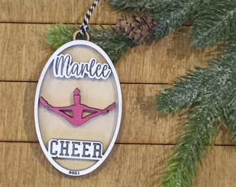 May include: A white oval wooden ornament with a pink silhouette of a cheerleader in a split position. The ornament has the name "Marlee" and the word "CHEER" with the year "2021" at the bottom.