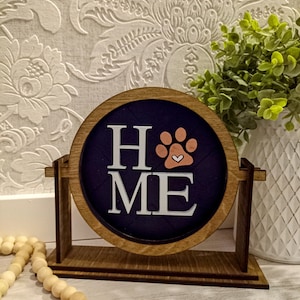 May include: A wooden sign with the word "HOME" in white letters and a paw print with a heart in the center. The paw print is peach colored and the heart is white. The sign is on a wooden stand and has a dark blue background.