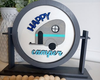 May include: A gray and white wooden sign with the words "Happy Camper" in blue and teal lettering. The sign features a cutout of a camper trailer with a teal stripe and a black wheel. The sign is on a gray wooden stand.