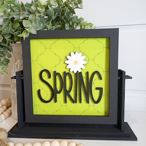 May include: A black framed sign with a green background and a white daisy. The sign says "SPRING" in black letters.