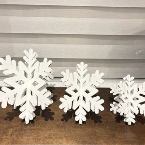 May include: Three white wooden snowflakes of varying sizes, arranged in a row on a wooden surface. The snowflakes are layered and have a 3D effect.