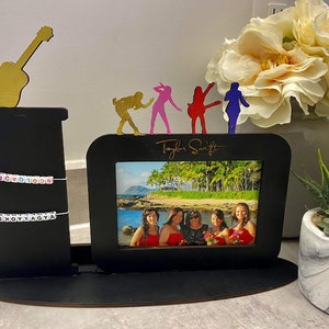 May include: Black wooden photo frame with a picture of five women. The frame has a guitar and dancing figures on top. The words "Taylor Swift" are written on the frame. Two beaded bracelets are strung across the black stand.