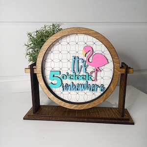 Puede incluir: Un letrero de madera con un flamenco rosa y el texto "It's 5 o'clock somewhere". El letrero está en un soporte de madera.