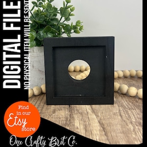 以下が含まれることがあります： 中央に丸い切り抜きのある黒い正方形の木製フレーム。フレームは木製の表面に置かれ、周りに白いビーズが散らばっています。フレームの下には「Find in our Etsy store」と「One Crafty Brit Co.」のテキストが表示されています。