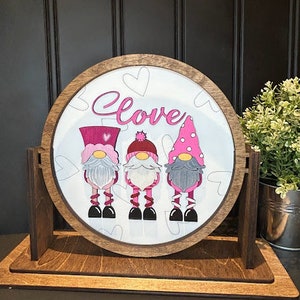 Könnte beinhalten: Ein rundes Holzschild mit weißem Hintergrund und dem Wort "Love" in Pink geschrieben. Drei Gnome sind auf dem Schild abgebildet, jeder mit einem rosa Hut und einem rosa-weißen Outfit. Das Schild steht auf einem Holzständer.
