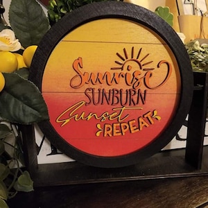 May include: A round wooden sign with a black frame and a sunset gradient background. The sign reads "Sunrise Sunburn Sunset Repeat" in a stylized font.