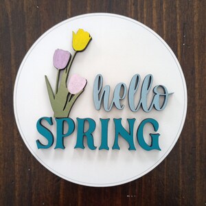 May include: A white circular wooden sign with the text "hello spring" in blue and teal lettering. The sign is decorated with three paper tulips in yellow, pink, and purple.