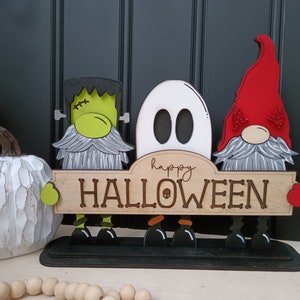 May include: Three Halloween gnomes, a green Frankenstein, a white ghost, and a red devil, stand in front of a wooden sign that reads "happy HALLOWEEN".