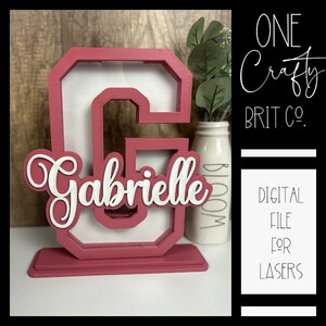 May include: A pink wooden letter G with a white cutout frame and the name "Gabrielle" in white lettering. The letter is on a pink base. The image is for a digital file for lasers.