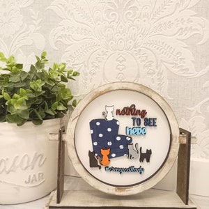 May include: A white wooden round sign with a blue and white polka dot armchair and five cats. The sign reads "nothing to see here #crazycatlady".