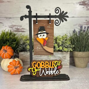 May include: Wooden Thanksgiving sign featuring a turkey wearing a pilgrim hat and the text "Gobble Til You Wobble".