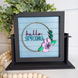 Op de afbeelding: Een zwart ingelijst bord met een blauwe achtergrond en witte strepen. Het bord zegt "hello spring" in een gouden cirkel met een roze bloem en groene bladeren.