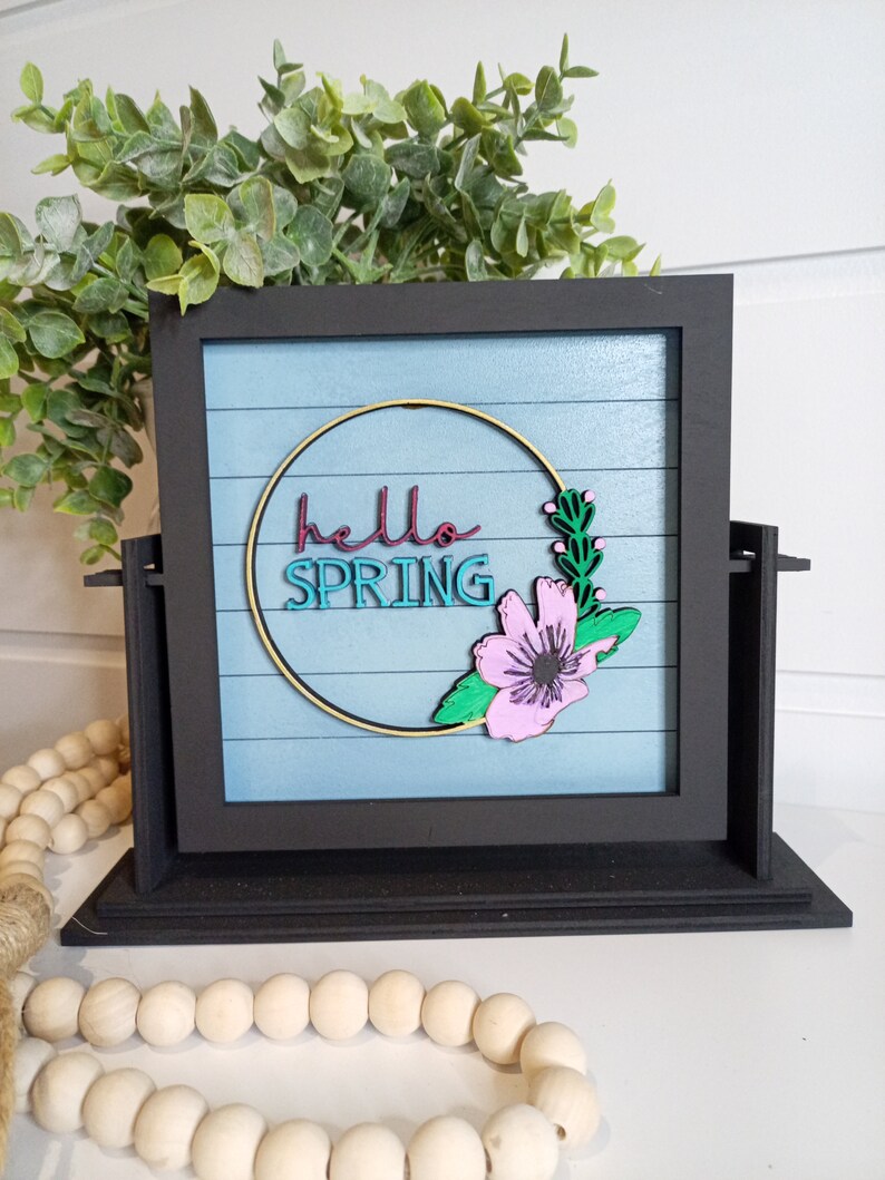 May include: A black framed sign with a blue background and white stripes. The sign says "hello spring" in a gold circle with a pink flower and green leaves.