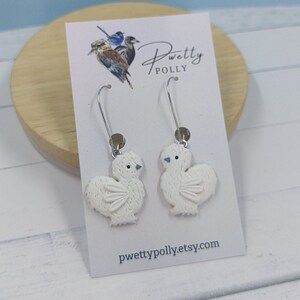 May include: White chicken dangle earrings with silver hooks. The earrings are handmade and feature a detailed design of a fluffy chicken. The earrings are displayed on a white card with the text "pwettypolly.etsy.com".
