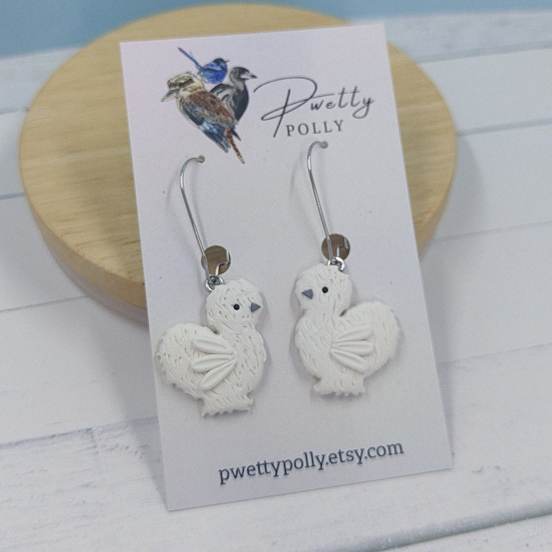 May include: White chicken dangle earrings with silver hooks. The earrings are handmade and feature a detailed design of a fluffy chicken. The earrings are displayed on a white card with the text "pwettypolly.etsy.com".