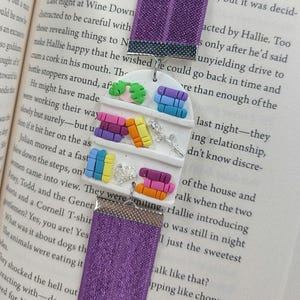 May include: A purple fabric bookmark with a white clay charm featuring miniature colourful books, a crown, scissors, and leaves. The bookmark is designed to be inserted into a book.