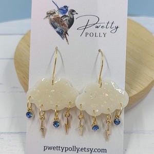 May include: Cloud-shaped earrings with gold hoops and lightning bolt charms. The earrings feature blue gemstone accents and are displayed on a card with the text "Pretty Polly" and the website pwettypolly.etsy.com.