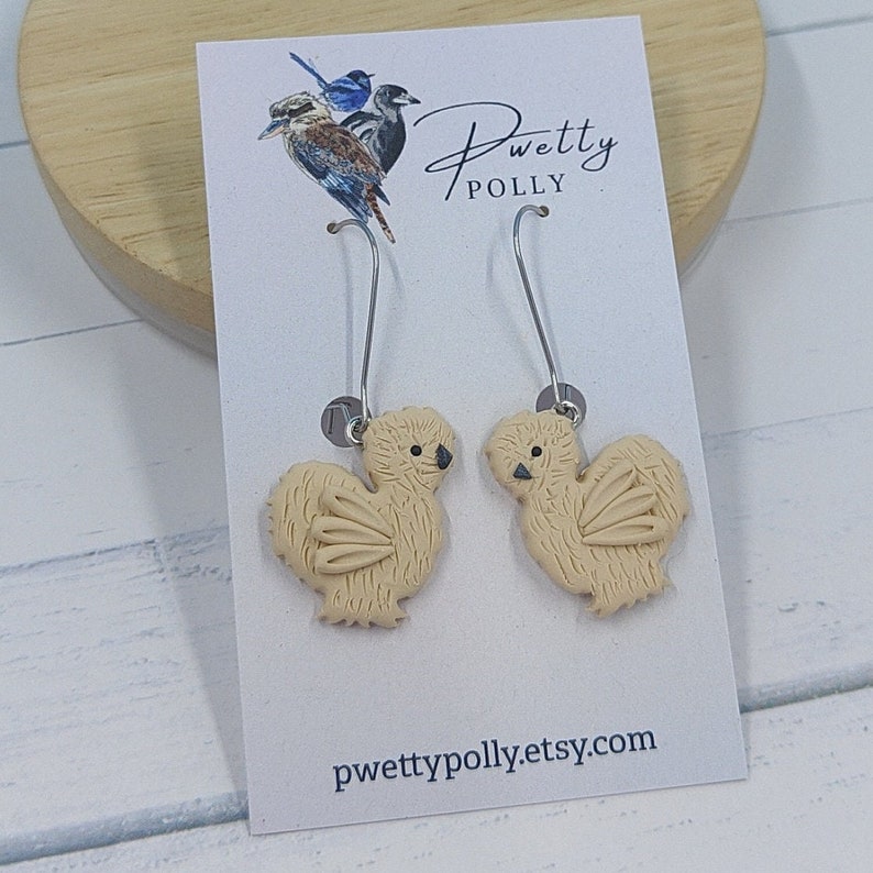 May include: A pair of dangle earrings featuring a beige, fluffy chicken design. The earrings are made of polymer clay and have silver hooks. The earrings are displayed on a white card with the text "pwettypolly.etsy.com".