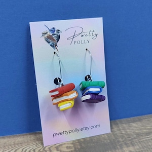 May include: Earrings with miniature rainbow-coloured book stacks. Each earring has three books on a silver hoop. The earrings are displayed on a white card with the text "Pretty Polly" and a website address.