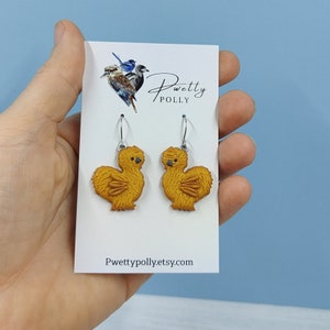 May include: A pair of gold-colored chicken earrings with silver hooks. The earrings are on a white card with the text "Pwetty Polly" and "Pwettypolly.etsy.com".