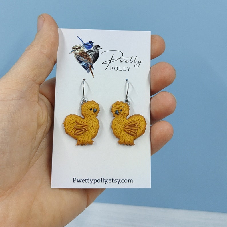 May include: A pair of gold-colored chicken earrings with silver hooks. The earrings are on a white card with the text "Pwetty Polly" and "Pwettypolly.etsy.com".