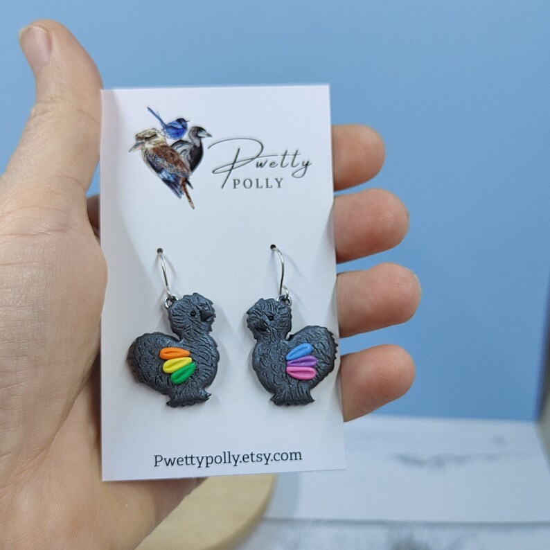 May include: A pair of black silkie chicken earrings with rainbow feathers. The earrings are on a white card with the text "Pwetty Polly" and "Pwettypolly.etsy.com".