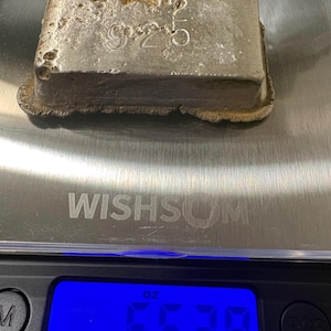 May include: A rectangular silver-colored metal ingot with the stamped number 925 on its top surface. The ingot is resting on a digital scale that displays a weight of 6.528 ounces. The metal has a rough, textured finish with visible pitting.