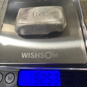 May include: A rectangular silver bar with the number 925 stamped on its surface, resting on a digital scale. The scale displays a weight of 9.253 ounces. The silver bar has a textured, metallic finish with rounded edges and a solid appearance.