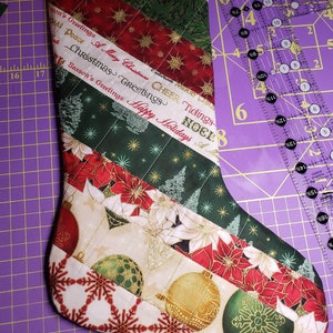 May include: A red and green Christmas stocking with a quilted design. The stocking is decorated with snowflakes, ornaments, and the words "Merry Christmas, Cheer Tidings, Noel, Happy Holidays".