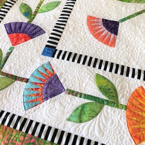 Pode incluir: Um edredom branco com uma borda listrada preta e branca. O edredom apresenta um padrão repetitivo de flores coloridas com folhas verdes. As flores são feitas de retalhos de tecido e têm um design único e moderno.