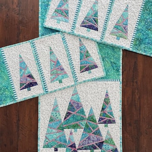 May include: A quilted table runner and two matching placemats featuring a patchwork design of stylized Christmas trees. The trees are constructed from various shades of blue, green, and purple fabric strips, set against a white background.