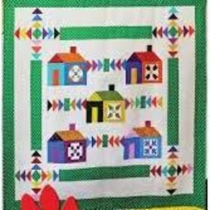 May include: A colorful quilt featuring a repeating pattern of houses in various colors, including orange, blue, and green. The houses are set against a white background with green borders and decorative geometric accents. The quilt is likely handmade.