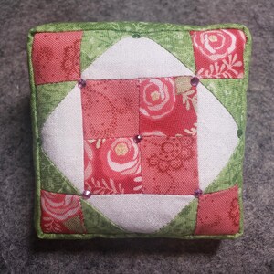 May include: A square pincushion with a patchwork design. The center features a white diamond shape surrounded by red squares with floral patterns. Green fabric with a leaf pattern borders the edges. Small purple gems are placed on the surface.
