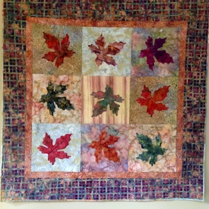 Peut inclure: Une courtepointe faite à la main avec neuf carrés, chacun avec une application de feuille colorée. Les feuilles sont dans des tons de rouge, orange et vert, sur un fond moucheté. La courtepointe a une bordure à motifs.