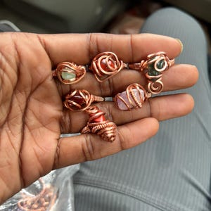 May include: A collection of handmade copper wire rings, each featuring a different gemstone. The rings showcase various colors, including green, red, and pink stones, all set within intricate wire designs. The rings are displayed on a hand.