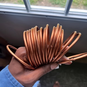 May include: A tightly coiled copper tube, held in a hand. The copper has a warm, reddish-brown hue and a smooth, reflective surface. The tubing is likely used for heat transfer or plumbing applications.