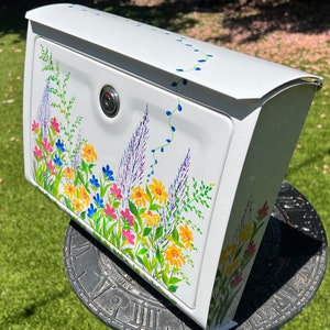 Op de afbeelding: Witte brievenbus met een bloemenontwerp. De brievenbus heeft een slot en is versierd met kleurrijke bloemen, waaronder gele, roze en blauwe bloemen, samen met paarse lavendel en groen blad. Het ontwerp is geschilderd op de voor- en zijkanten.