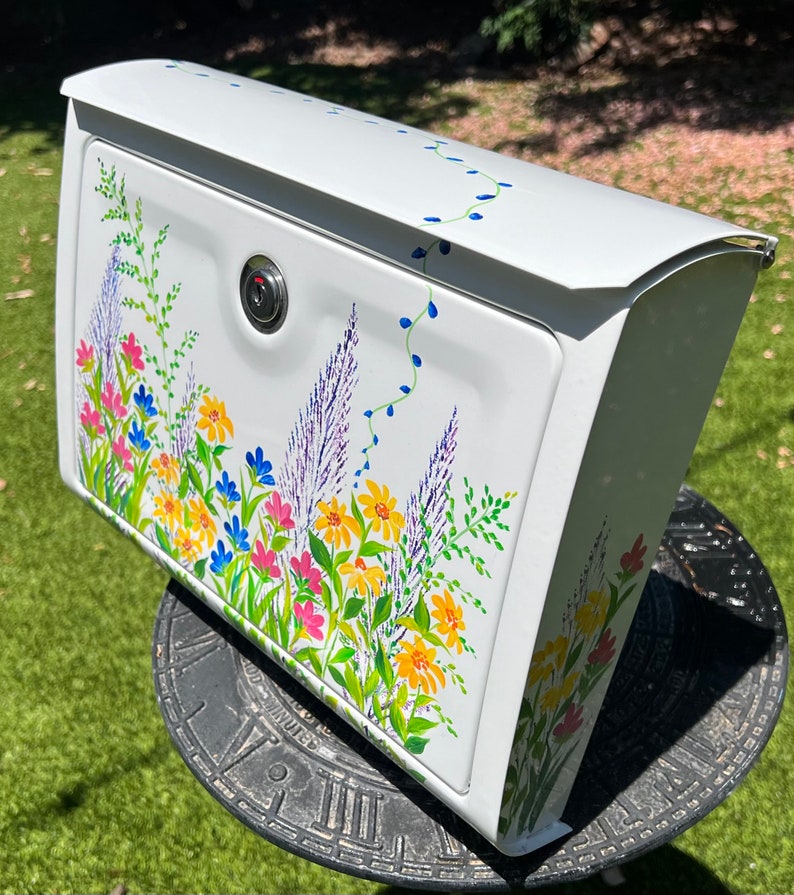 Op de afbeelding: Witte brievenbus met een bloemenontwerp. De brievenbus heeft een slot en is versierd met kleurrijke bloemen, waaronder gele, roze en blauwe bloemen, samen met paarse lavendel en groen blad. Het ontwerp is geschilderd op de voor- en zijkanten.