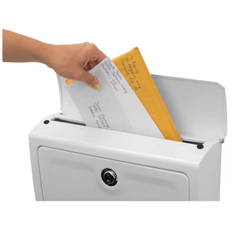 Op de afbeelding: Een witte brievenbus met een scharnierend deksel, een slot en een gleuf voor post. Een hand steekt een witte envelop en een bruine envelop in de gleuf. De enveloppen hebben handgeschreven adressen.