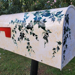 Può includere: Una cassetta postale bianca con un disegno dipinto di edera verde e blu. La cassetta postale ha una bandiera rossa ed è montata su un palo nero.