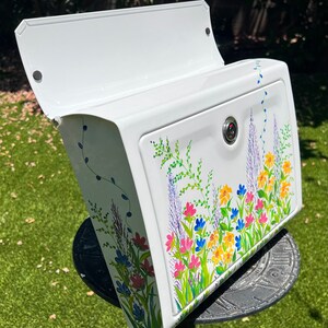 Op de afbeelding: Witte brievenbus met een handgeschilderd bloemmotief. Het ontwerp omvat roze, blauwe, gele en paarse bloemen met groene bladeren. De brievenbus heeft een scharnierend deksel en een slot.