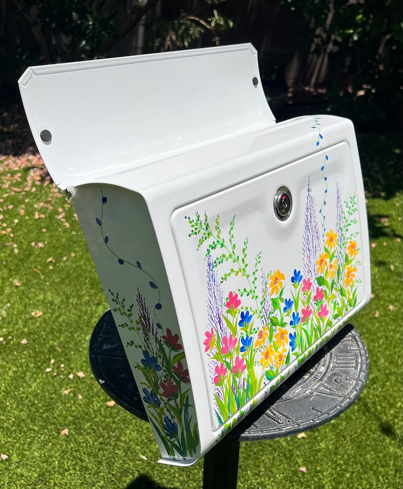 Op de afbeelding: Witte brievenbus met een handgeschilderd bloemmotief. Het ontwerp omvat roze, blauwe, gele en paarse bloemen met groene bladeren. De brievenbus heeft een scharnierend deksel en een slot.
