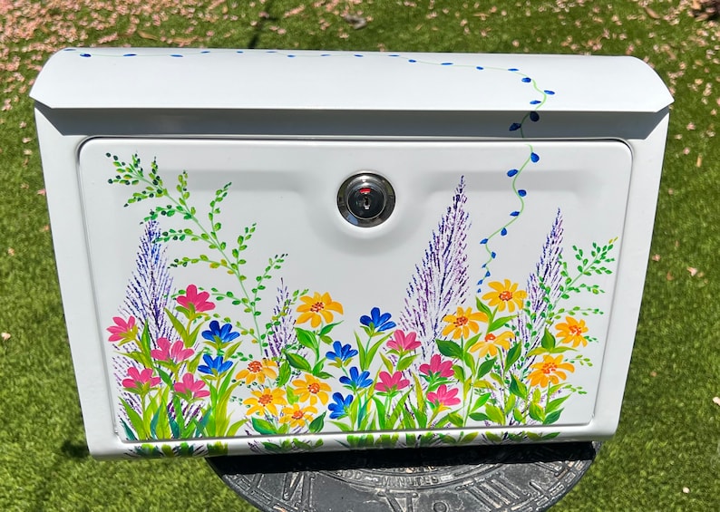 Op de afbeelding: Witte metalen brievenbus met een bloemenontwerp. De brievenbus heeft een slot en is versierd met kleurrijke bloemen in roze, geel en blauw, samen met groene bladeren. Blauwe wijnstokaccenten voegen een grillige toets toe.