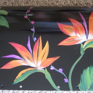 May include: A black mailbox with a hand-painted design of tropical flowers and leaves. The flowers are orange, yellow, white, and purple. The leaves are green.
